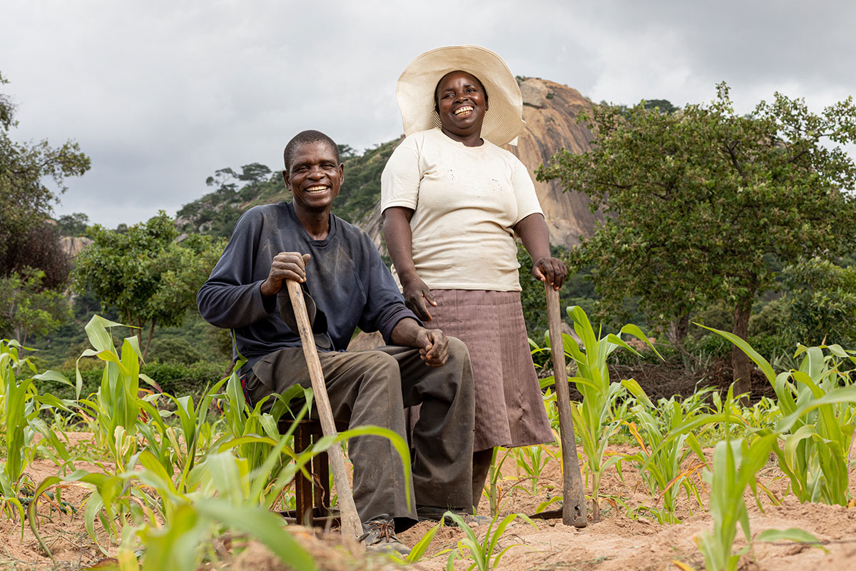 A standing woman and sitting man pose next to each other in a field. They are both smiling widely and leaning on wooden sticks.